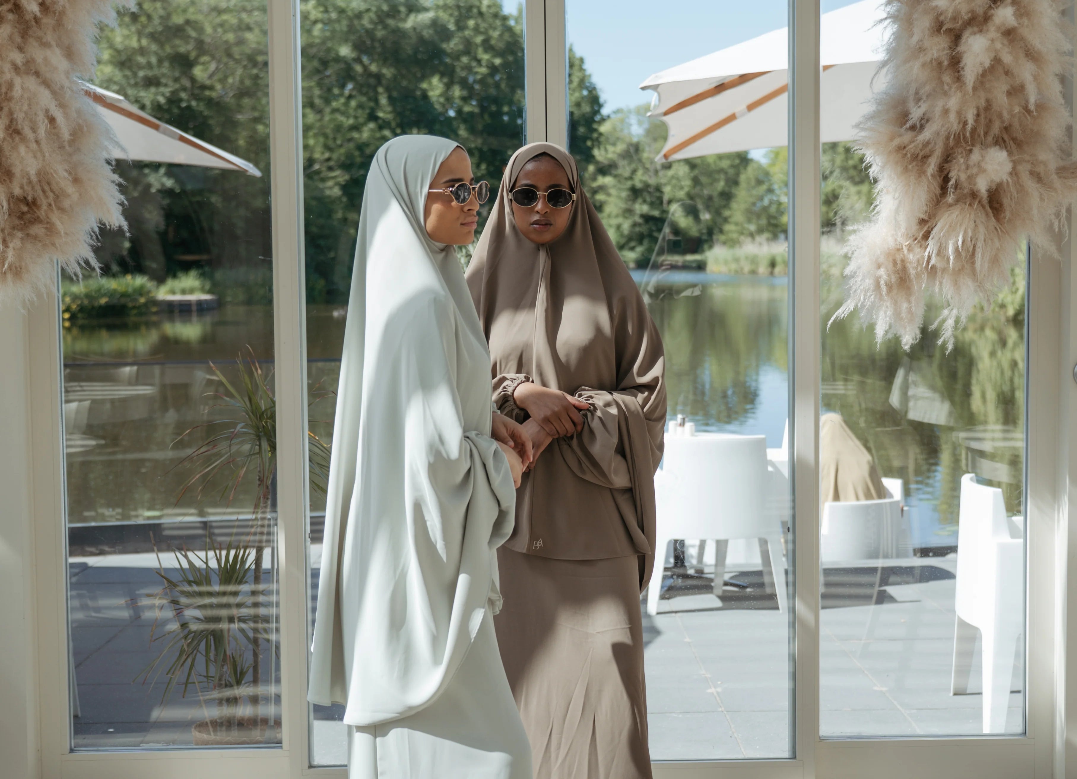 Twee vrouwen in elegante, losvallende hijabs staan binnen bij een glazen wand met uitzicht op een groen landschap. Ze dragen beiden zonnebrillen; de vrouw links draagt een lichtgrijze outfit, de vrouw rechts een taupekleurige. Ze staan rustig tegenover elkaar en houden elkaars hand lichtjes vast.